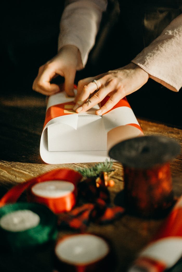 Close-up of a person wrapping a Christmas gift with red ribbon on a wooden table.